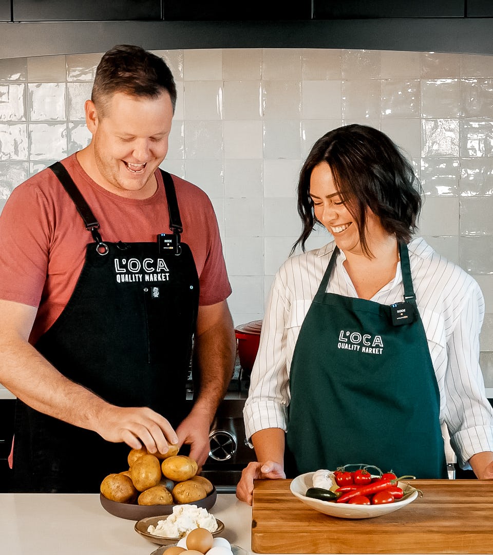 Two employees in black L'OCA aprons are smiling and chopping potatoes and other produce in a kitchen.
