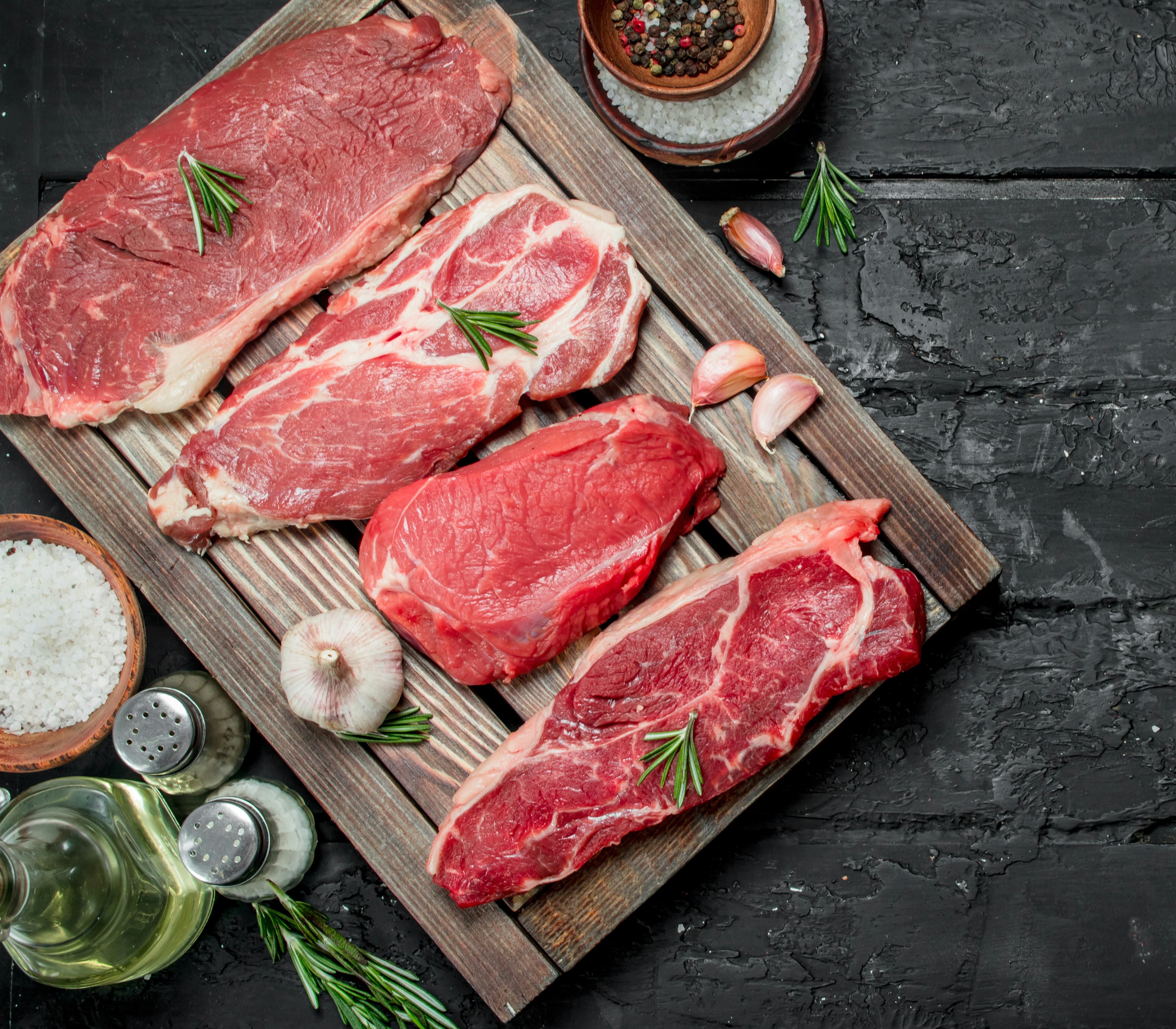 Four cuts of uncooked meat are resting on a wooden board. Salt, pepper and other seasonings are next to the board.