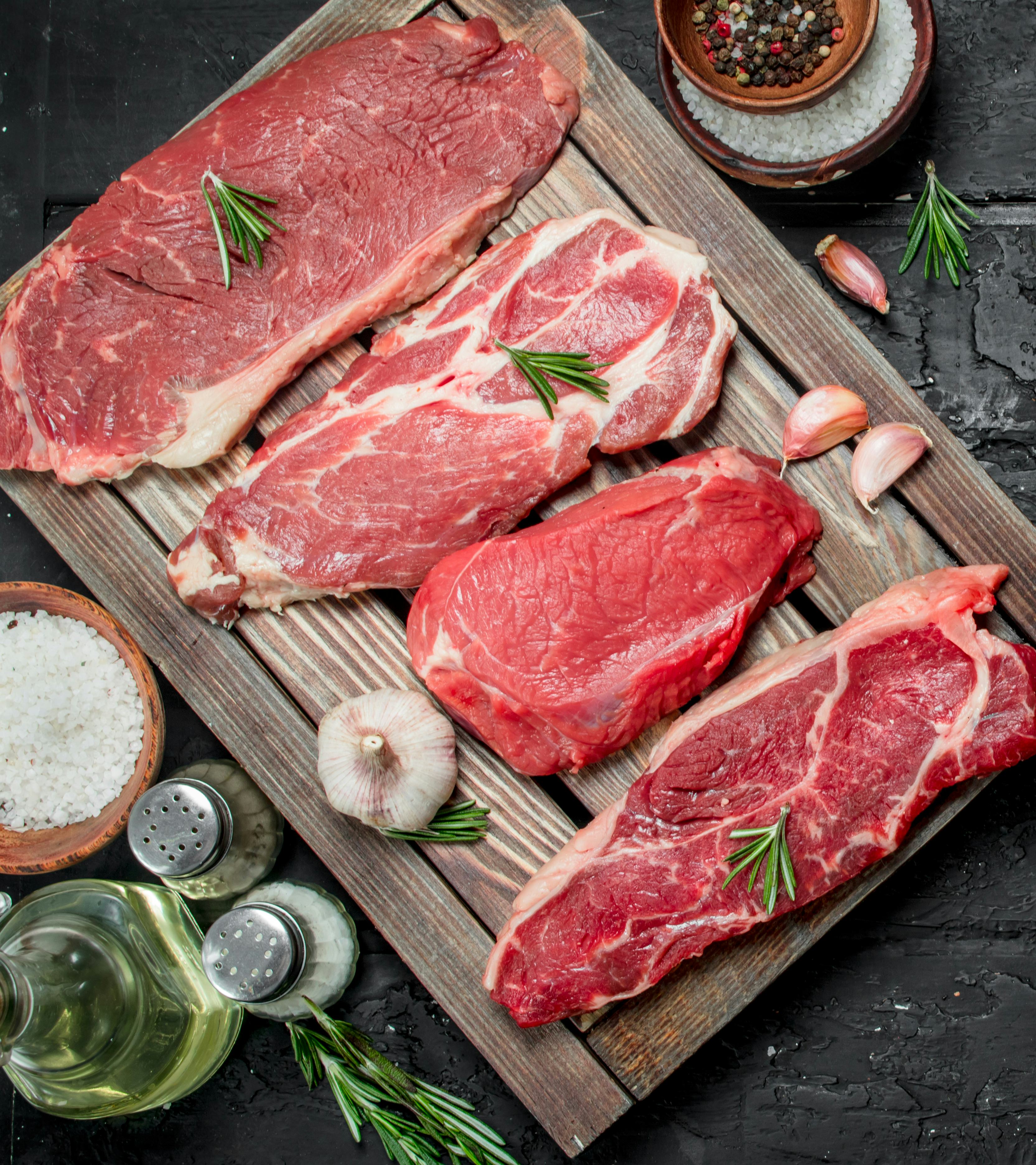 Four cuts of uncooked meat are resting on a wooden board. Salt, pepper and other seasonings are next to the board.