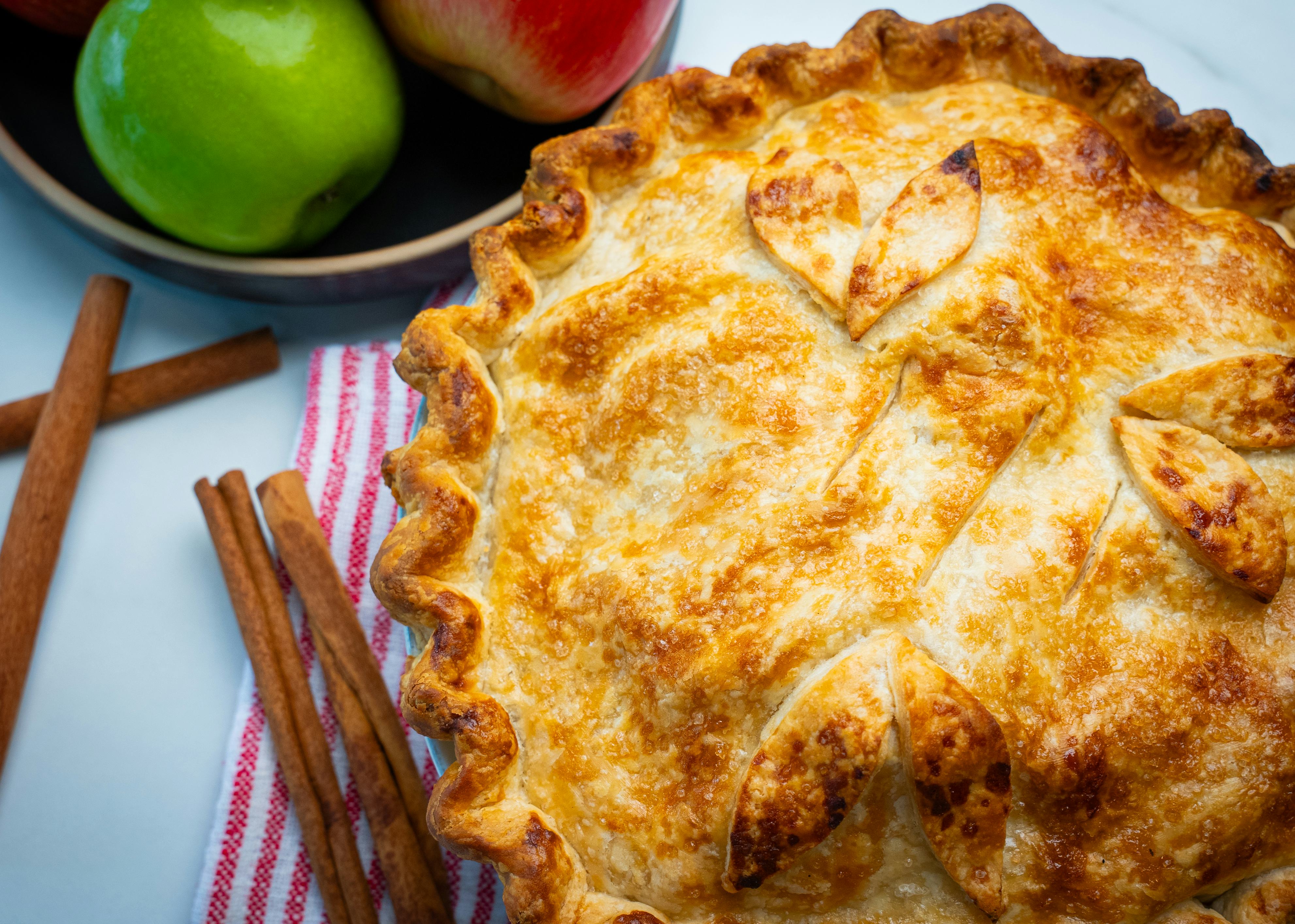 An apple pie with decorated pastry. Next to it are a few cinnamon sticks, and a bowl with two apples.