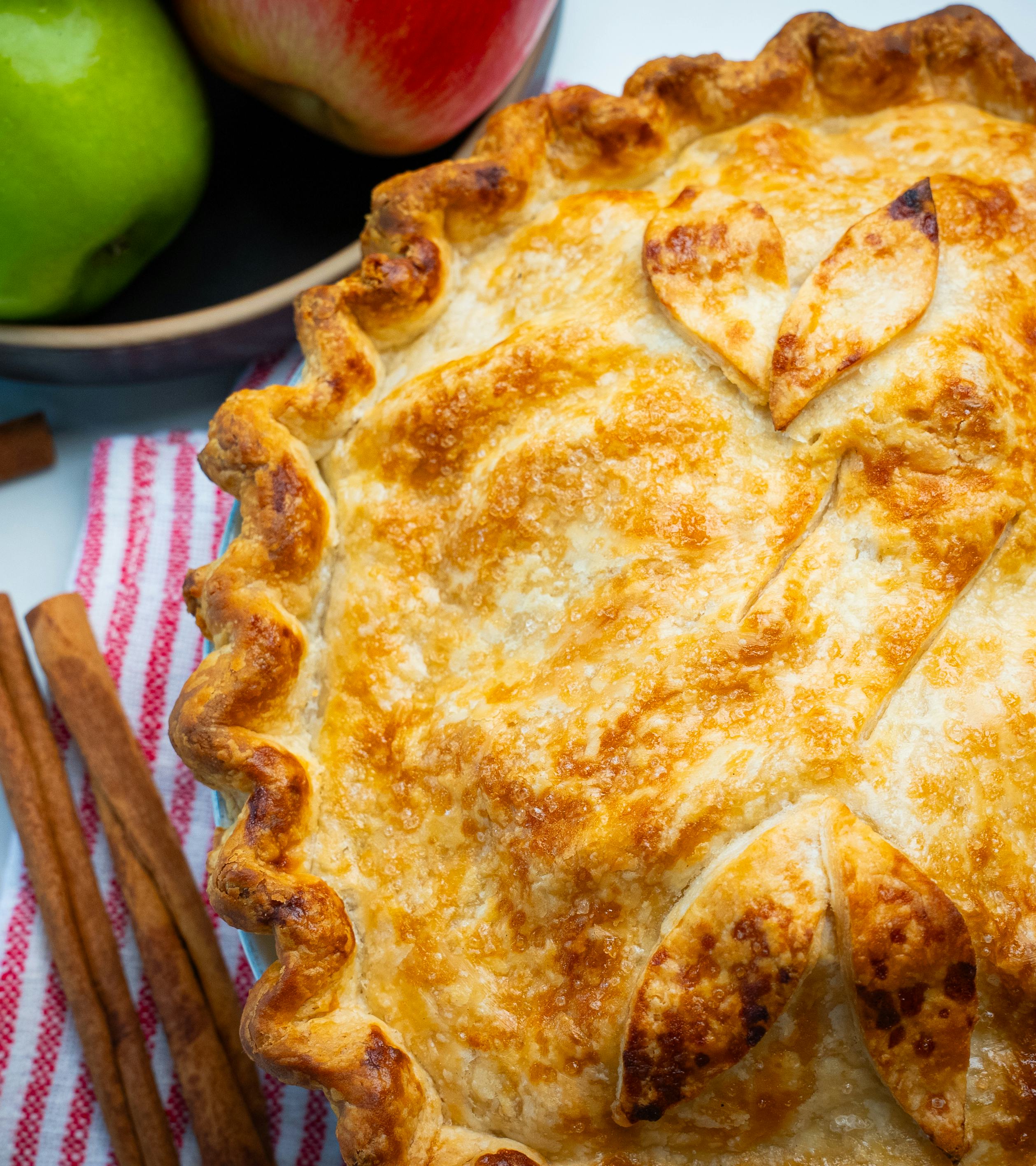 An apple pie with decorated pastry. Next to it are a few cinnamon sticks, and a bowl with two apples.